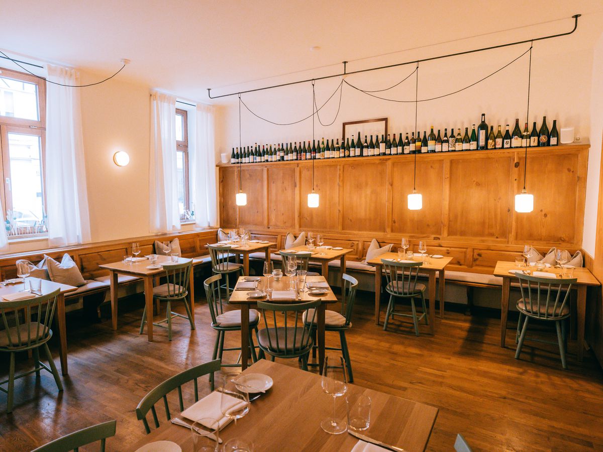 A restaurant interior with bright wooden floors, tables, and an accent wall lined with bottles. 