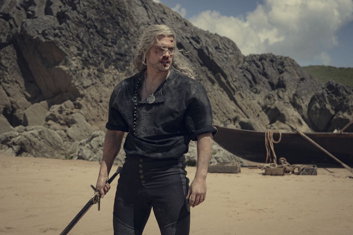 Geralt (Henry Cavill) with cuts on his face standing on a beach holding a sword