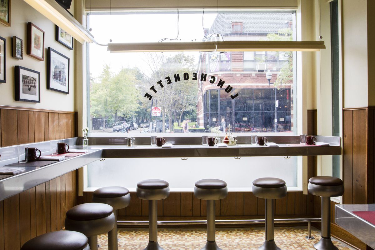 A wood paneled diner with a wooden countertop and built-in stools. The front counter is built up against a picture window that looks out onto a city street.