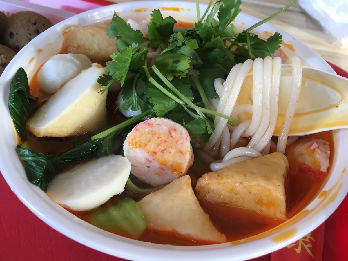 Fish balls, springy rice noodles, and greens fill a soup with a reddish-pink broth in a white plastic takeout bowl.
