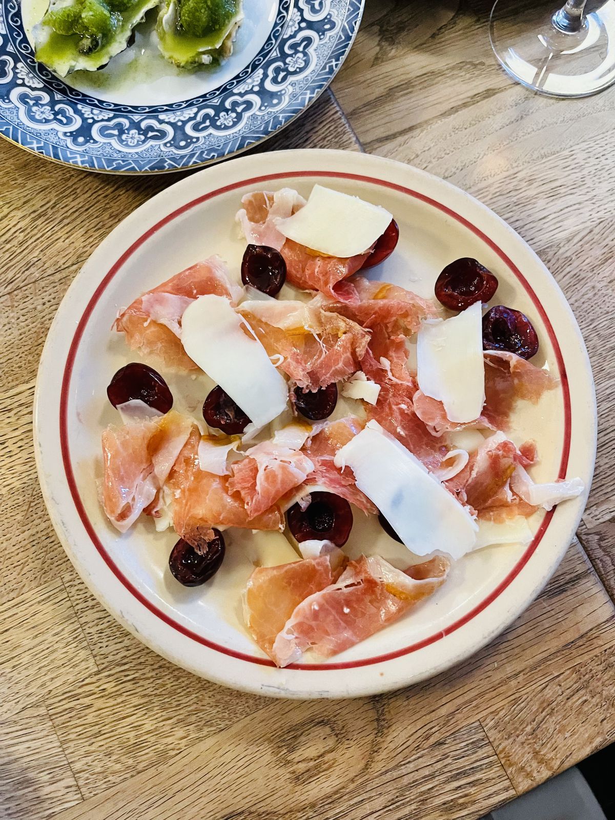 A plate of thinly shaved ham slices, cherries and shaved cheese on a table.