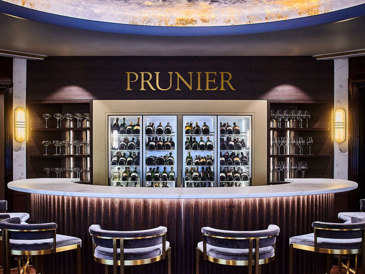 A glitzy, gold-accented round bar with a wall of Champagne fridges, a cloud-like sky, midcentury bar stools, and the name Prunier in large letters. 