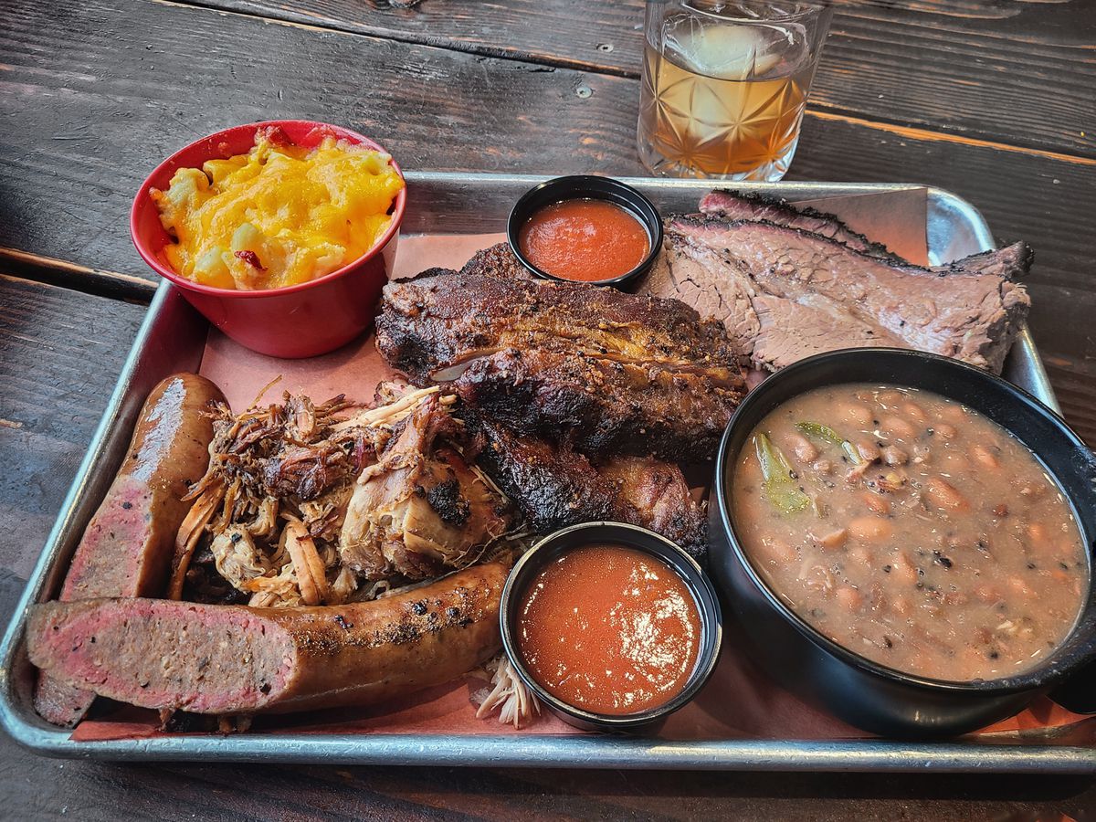 Sausage, pulled pork, brisket, sauces and beans are shown with mac and cheese on a metal tray on top of a wooden table.