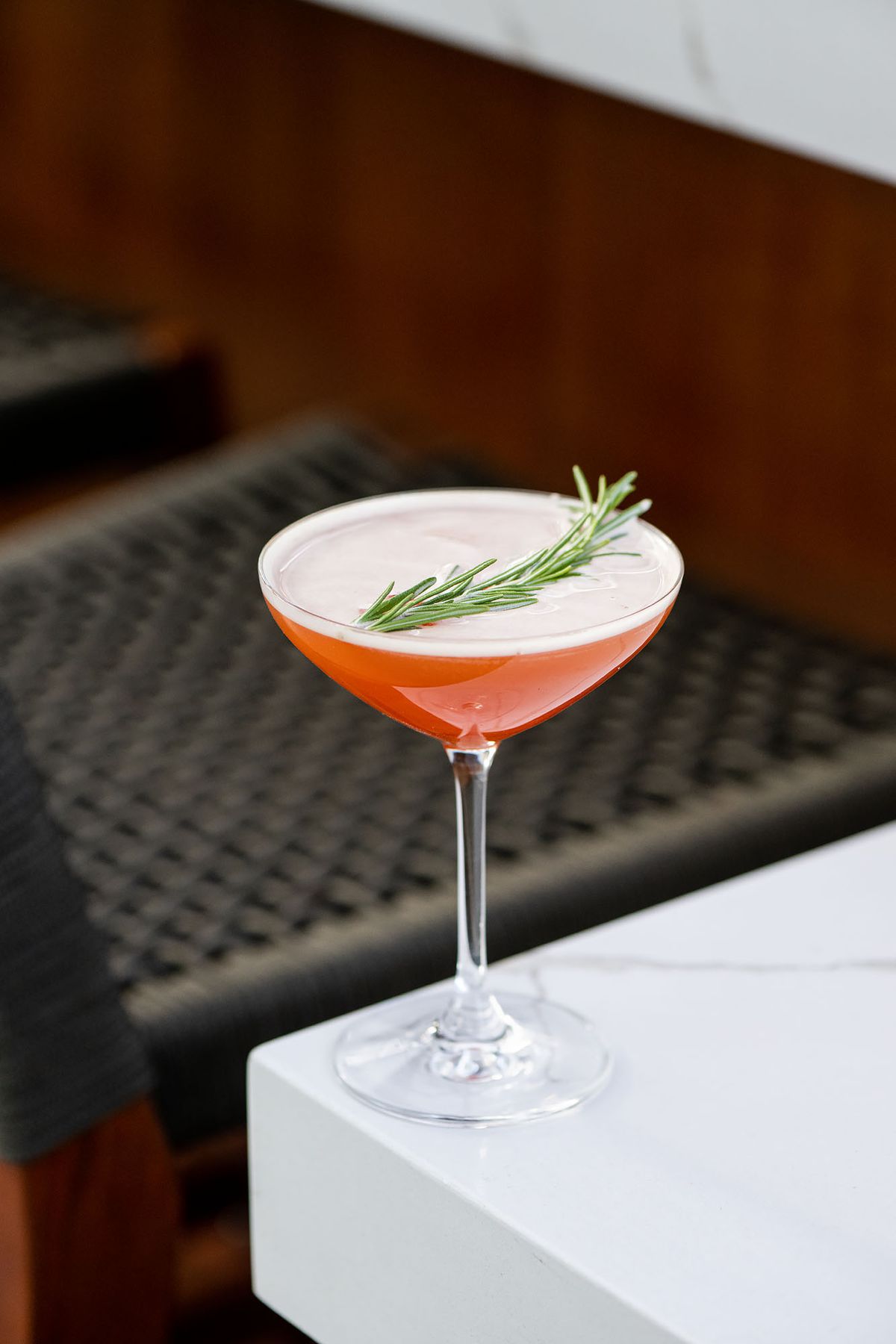 An orange-pink cocktail with a rosemary sprig on top.