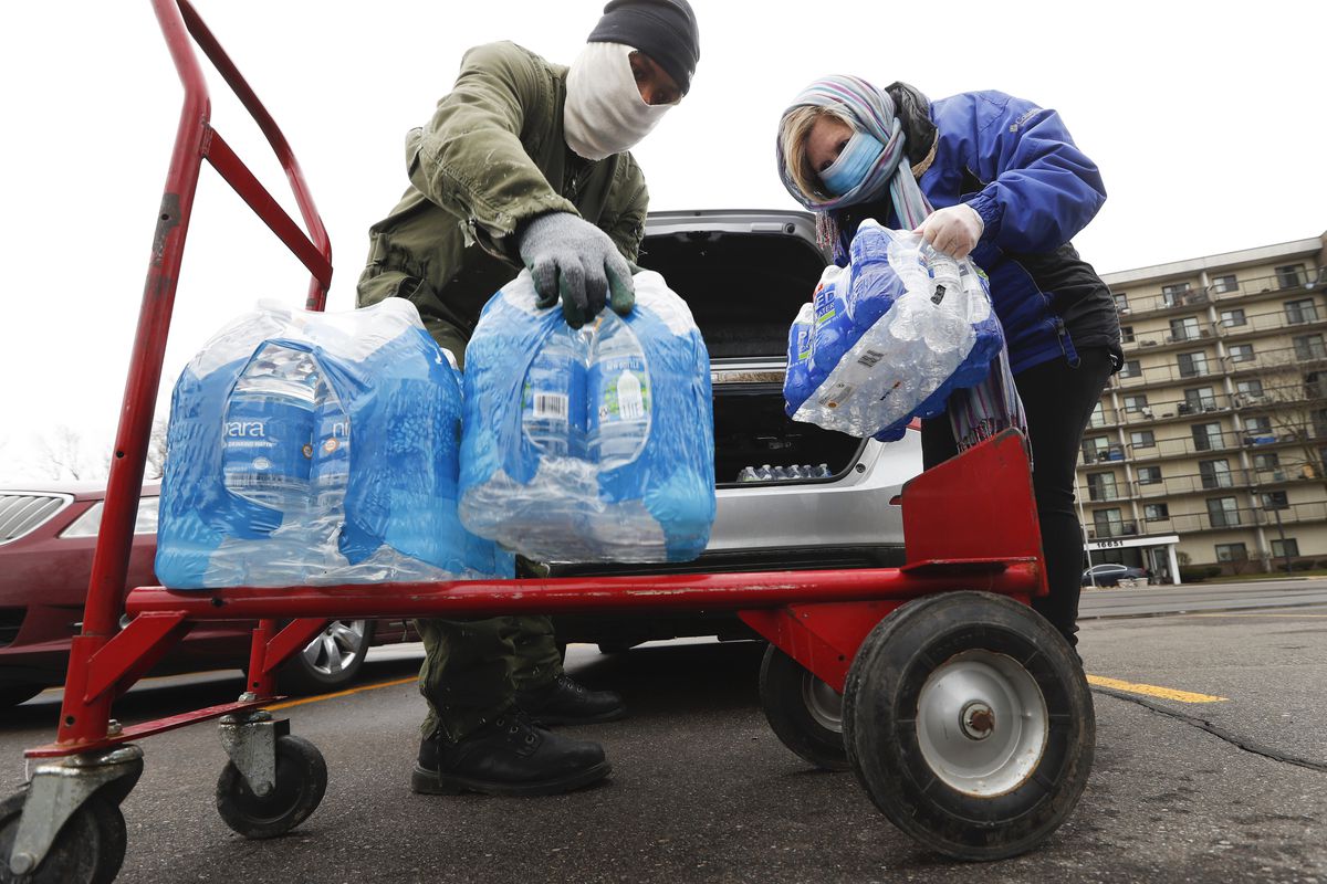 Two people wearing face masks place cases of bottled water from a van onto a dolly.