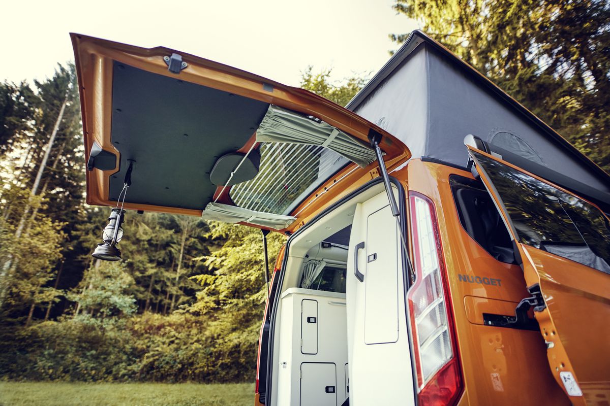 An orange camper van with the rear hatch door open. There are trees in the background. 