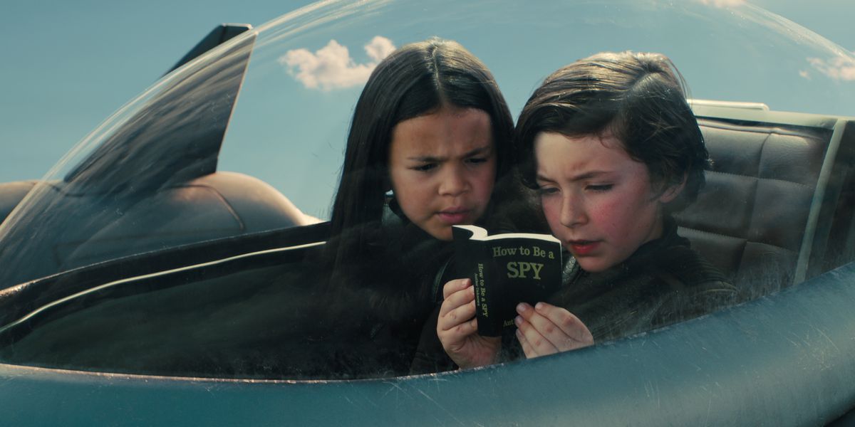 Everly Carganilla as Patty Torrez and Connor Esterson as Tony Torrez inside the cockpit of a personal stealth jet reading a small book titled “How To Be A Spy” in Spy Kids: Armageddon.