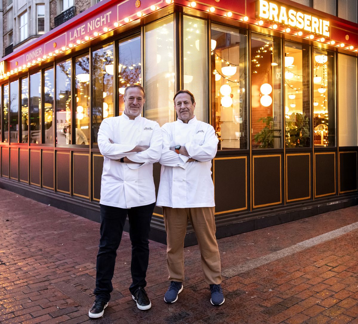 Two men in white chefs coats stand outside their Blue Ribbon Brasserie restaurant at dusk.
