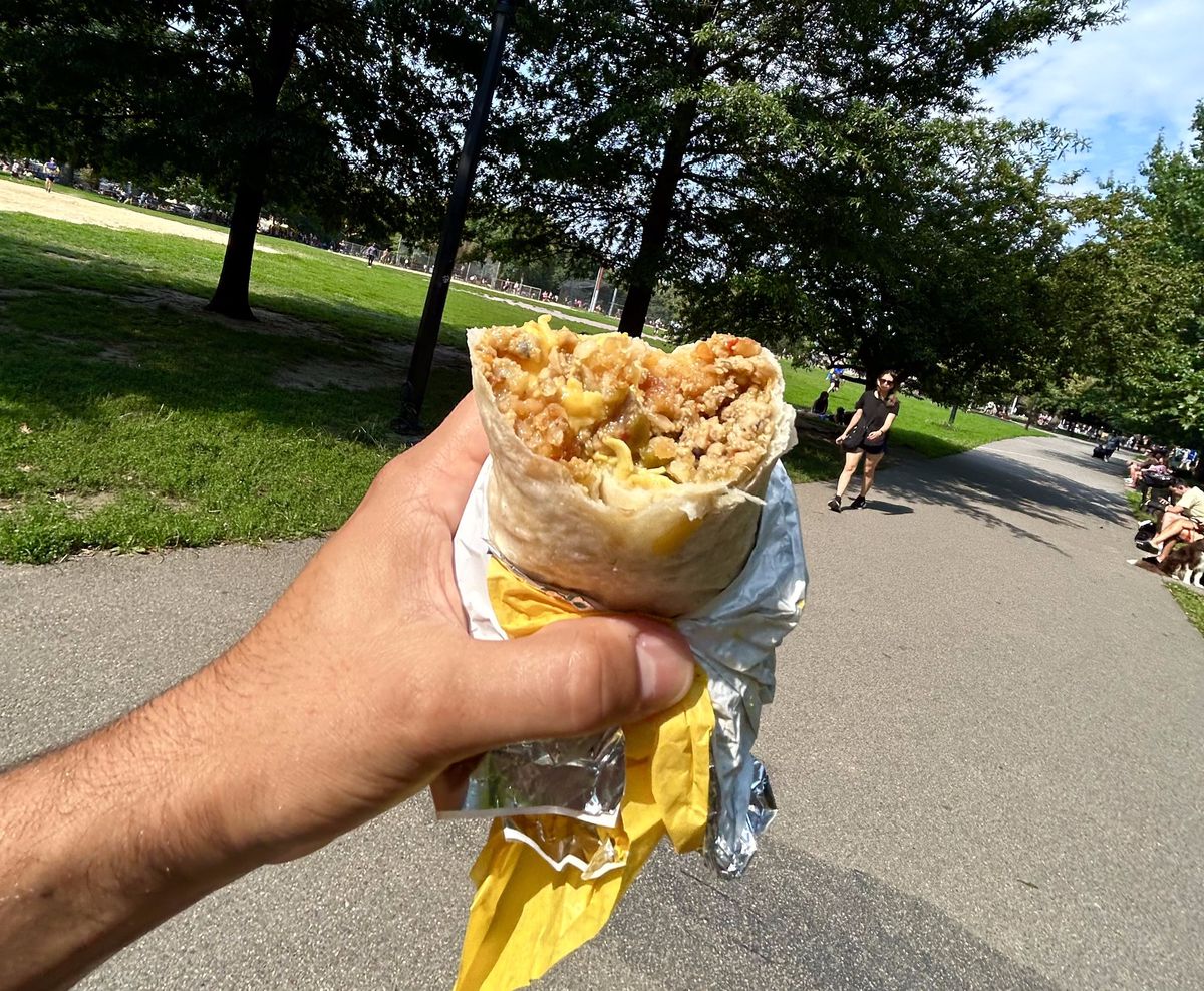 A hand holds a breakfast burrito for a photograph in a park.