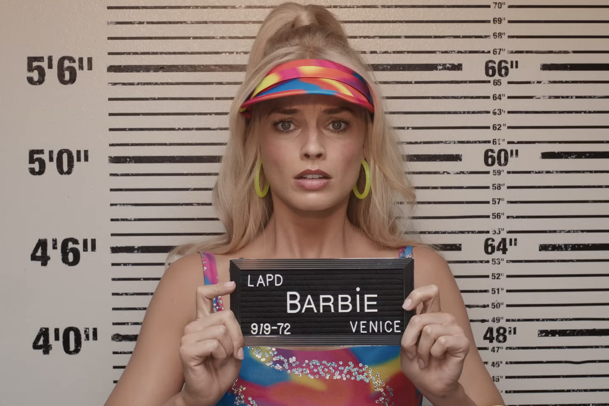 A still from the Barbie movie that shows Barbie taking a mugshot. Barbie is played by Margot Robbie and she looks distressed as she holds up a sign that says Barbie LAPD Venice.