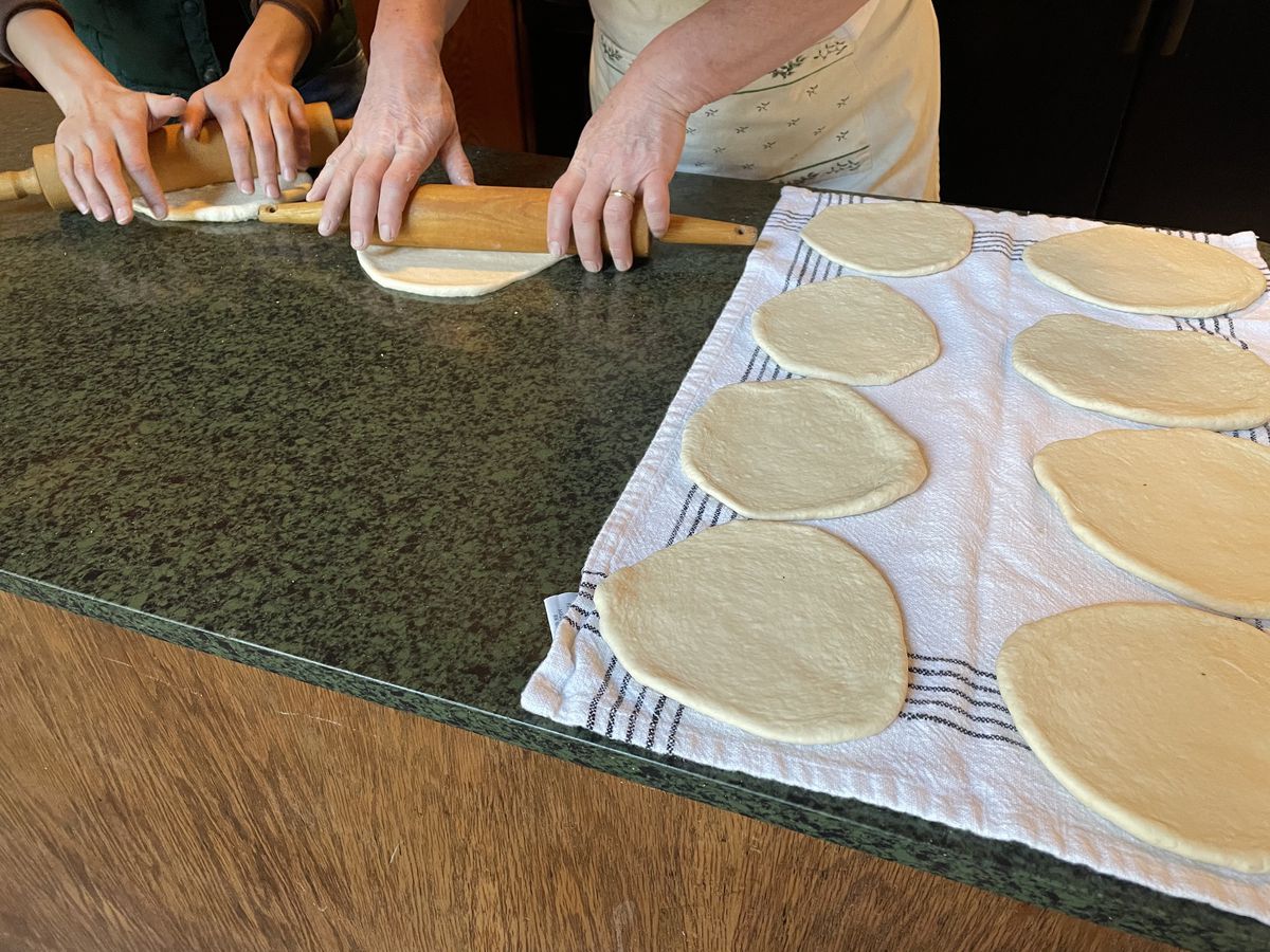 Two hands using rolling pins to roll out manikish dough into circles on a countertop, with eight already rolled on a white towel with blue stripes. 
