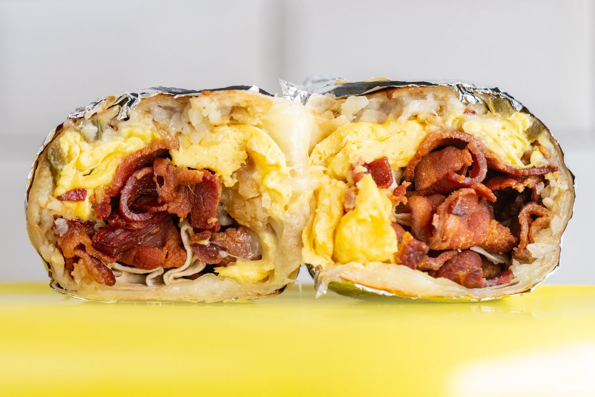 A close up shot of a cut interior of a breakfast burrito with eggs, potatoes, and bacon on a yellow table.