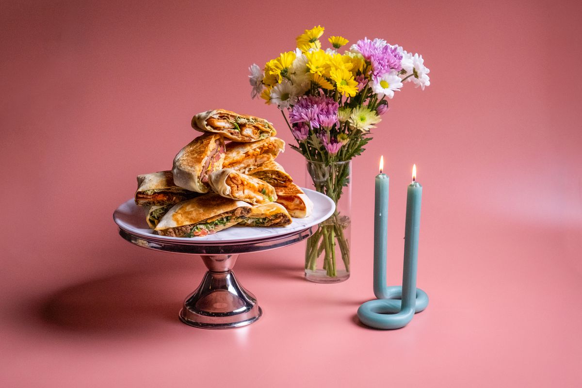 A plate of tortilla-wrapped pockets of meat and cheese on a silver tray next to a candle and a bouquet of flowers on a pink background. 