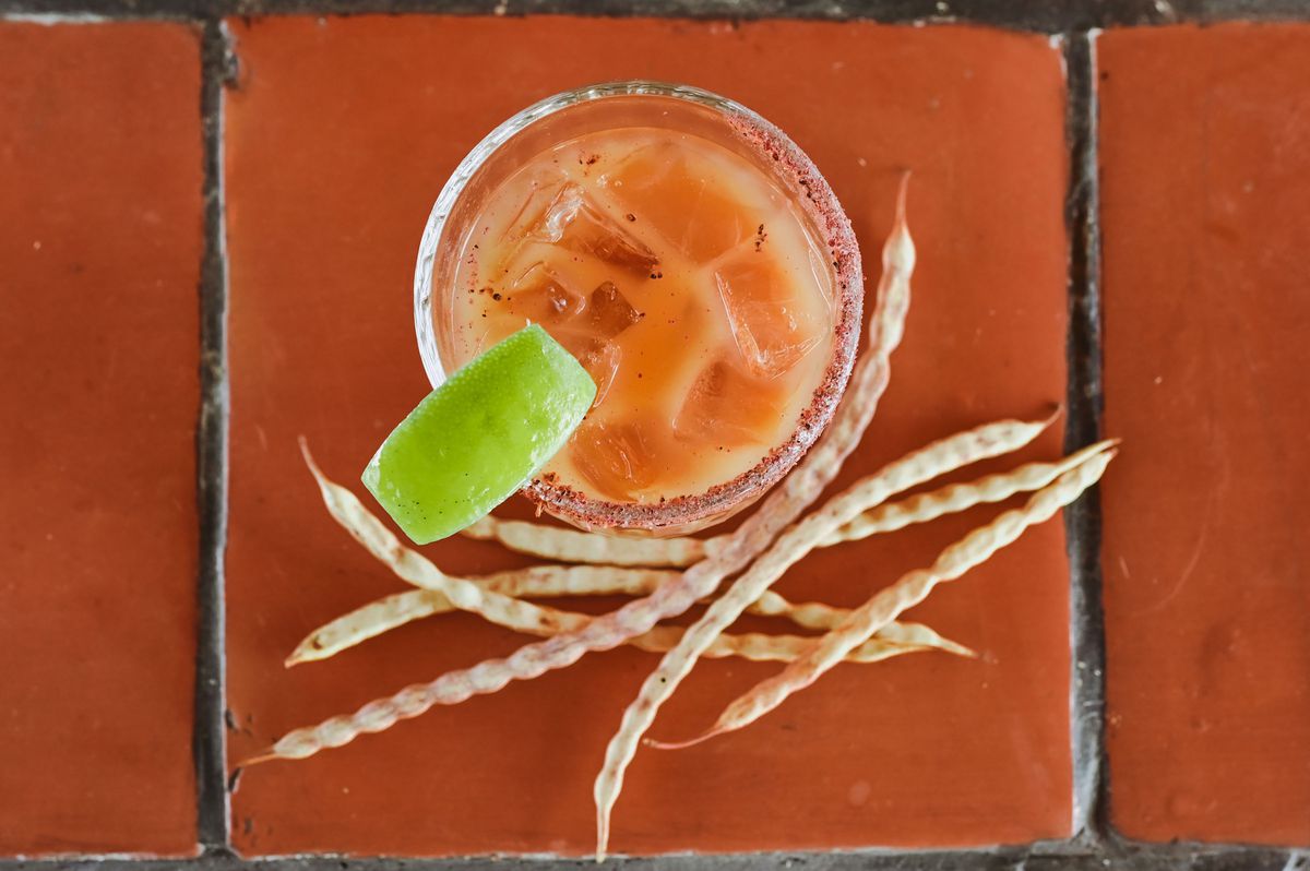 A pale pink-orange cocktail shot from above with a lime slice next to dried-out pods on a pale pink tiled counter.