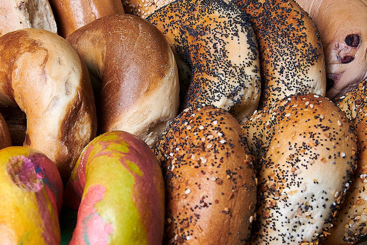 A closeup of many bagels stood on their edges and lined up close together, including a rainbow bagel and some with seeds.