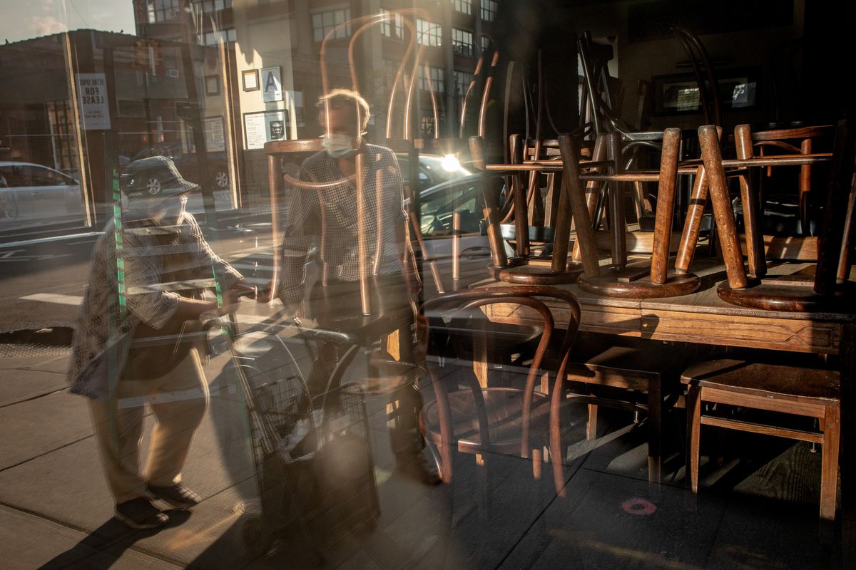 A window looking into a closed restaurants, where stools are on top of a table and a man with a mask is in the reflection.