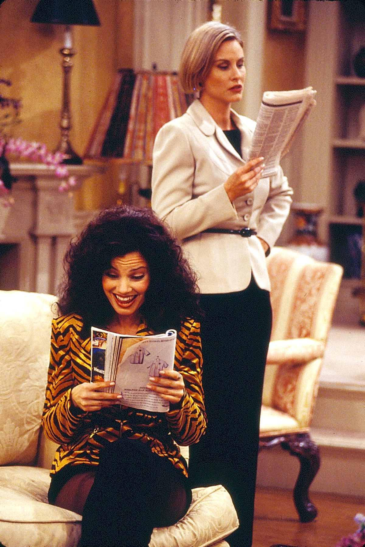 On the set of The Nanny, Fran Drescher (as Fran Fine) sits on the couch and reads a magazine, while Lauren Lane (as Sissy Babcock) stands up and holds a newspaper.