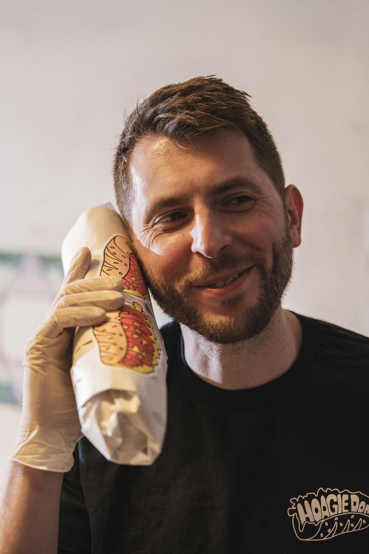 A man with a beard and wearing a food safety glove holds a hoagie wrapped in paper up to his ear like it’s a telephone.