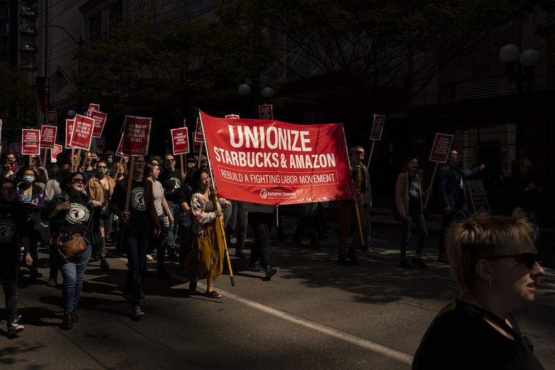 A crowd of people march down a street carrying signs and banners that read “Unionize Starbucks and Amazon.”