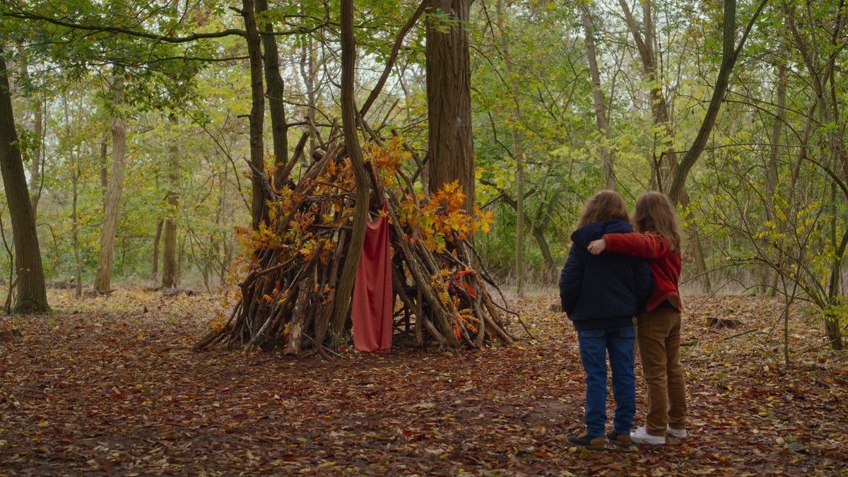 Two girls in a forest with their backs to the camera, hugging each other and looking at a leaf-covered tree shelter they built in Petite Maman