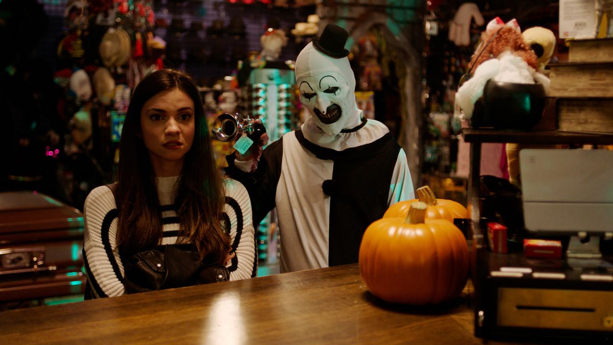 Lauren LaVera as Sienna Shaw stands uncomfortably at a Halloween-store counter, staring forward as David Howard Thornton honks a little silver bicycle horn in her ear as grinning killer mime Art the Clown in Terrifier 2