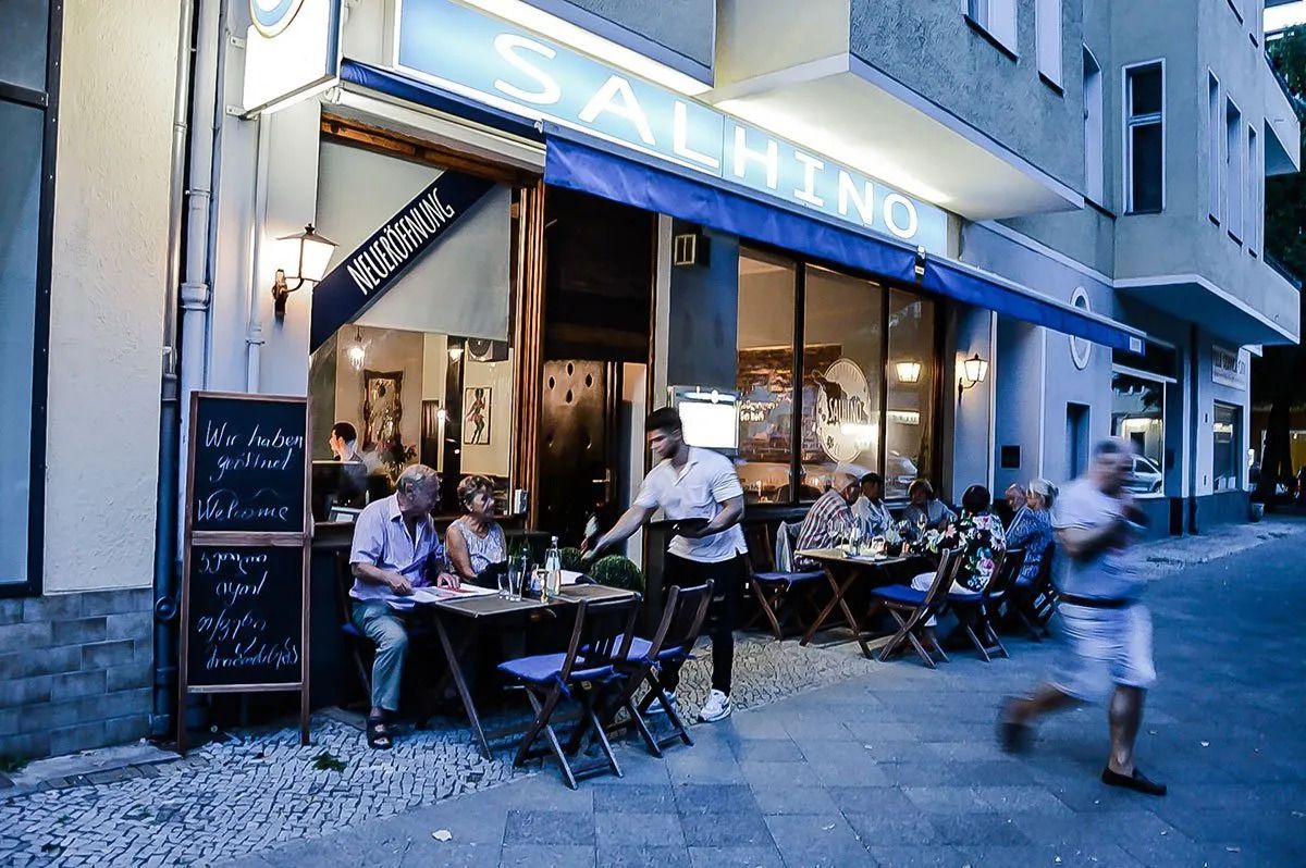 A restaurant exterior with diners seated at small picnic tables on a cobblestone street