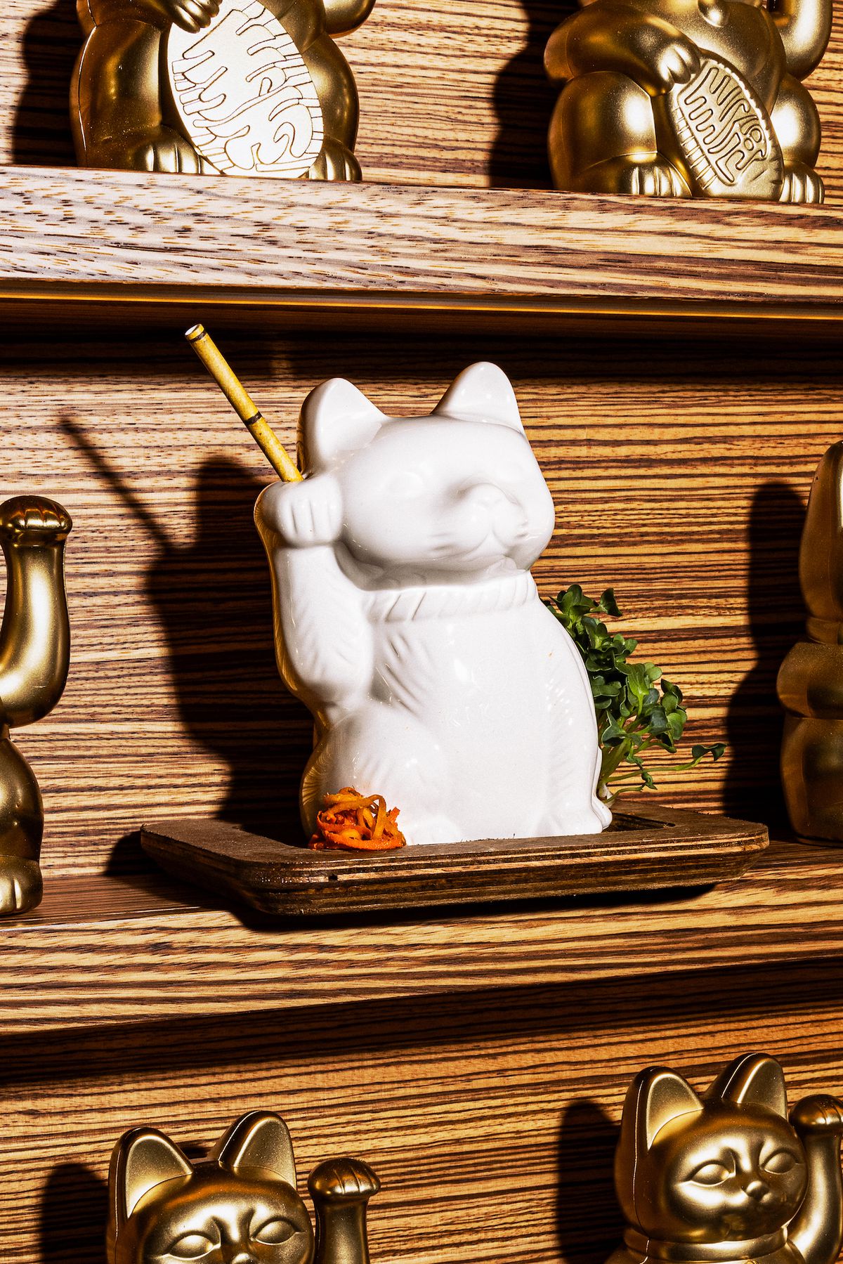 A white porcelain drinking vessel on a wall with a waiving cat arm and golden straw at new LA restaurant Lucky Mizu.