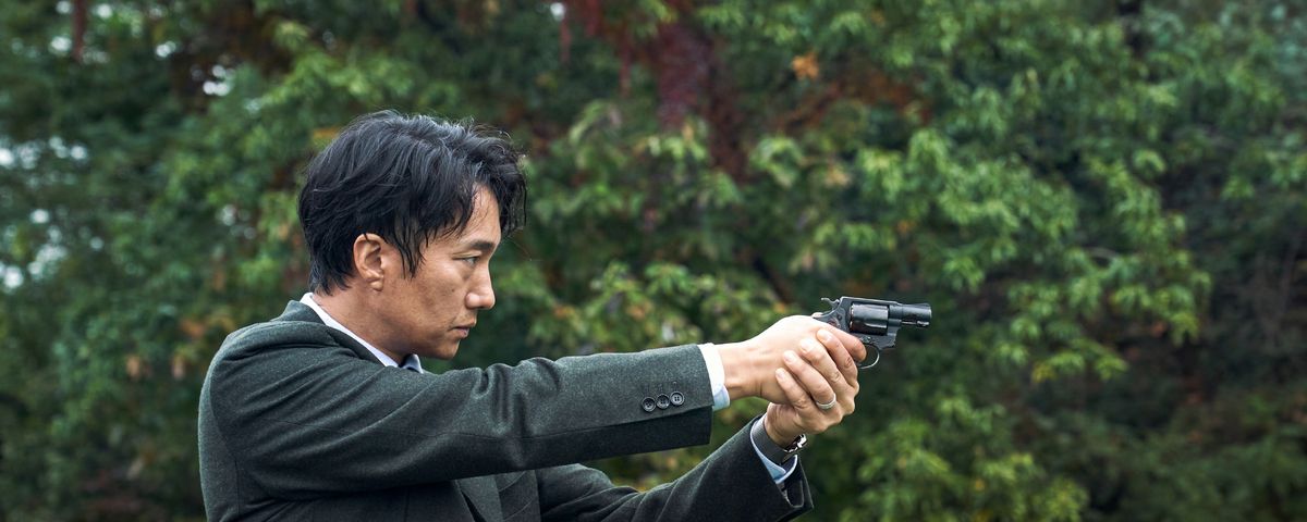 Detective Hae-jun (Park Hae-il), in a dapper suit, points a gun off screen against a backdrop of dark greenery in Decision to Leave