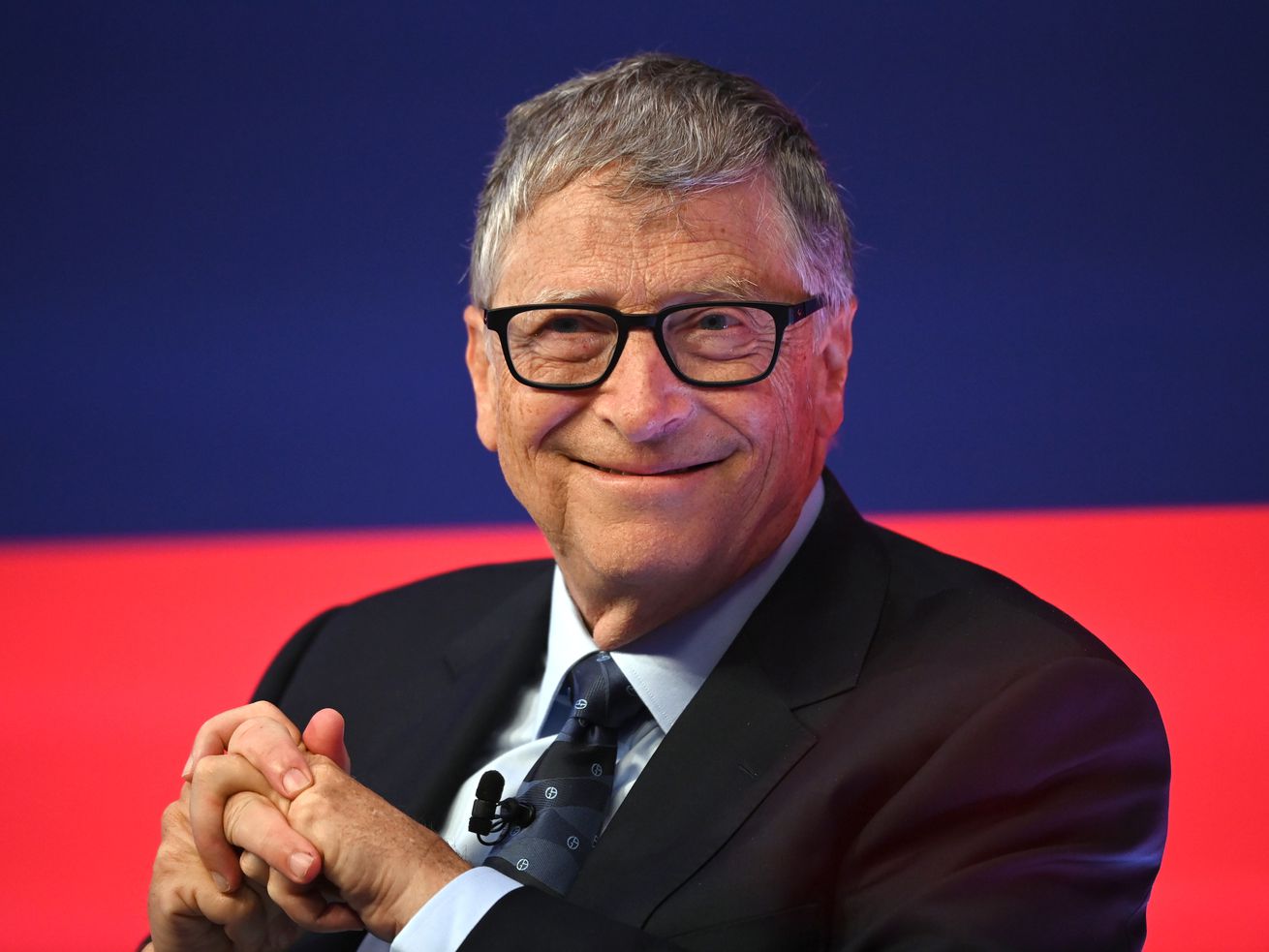 Bill Gates smiling and sitting onstage in front of a red and blue background.