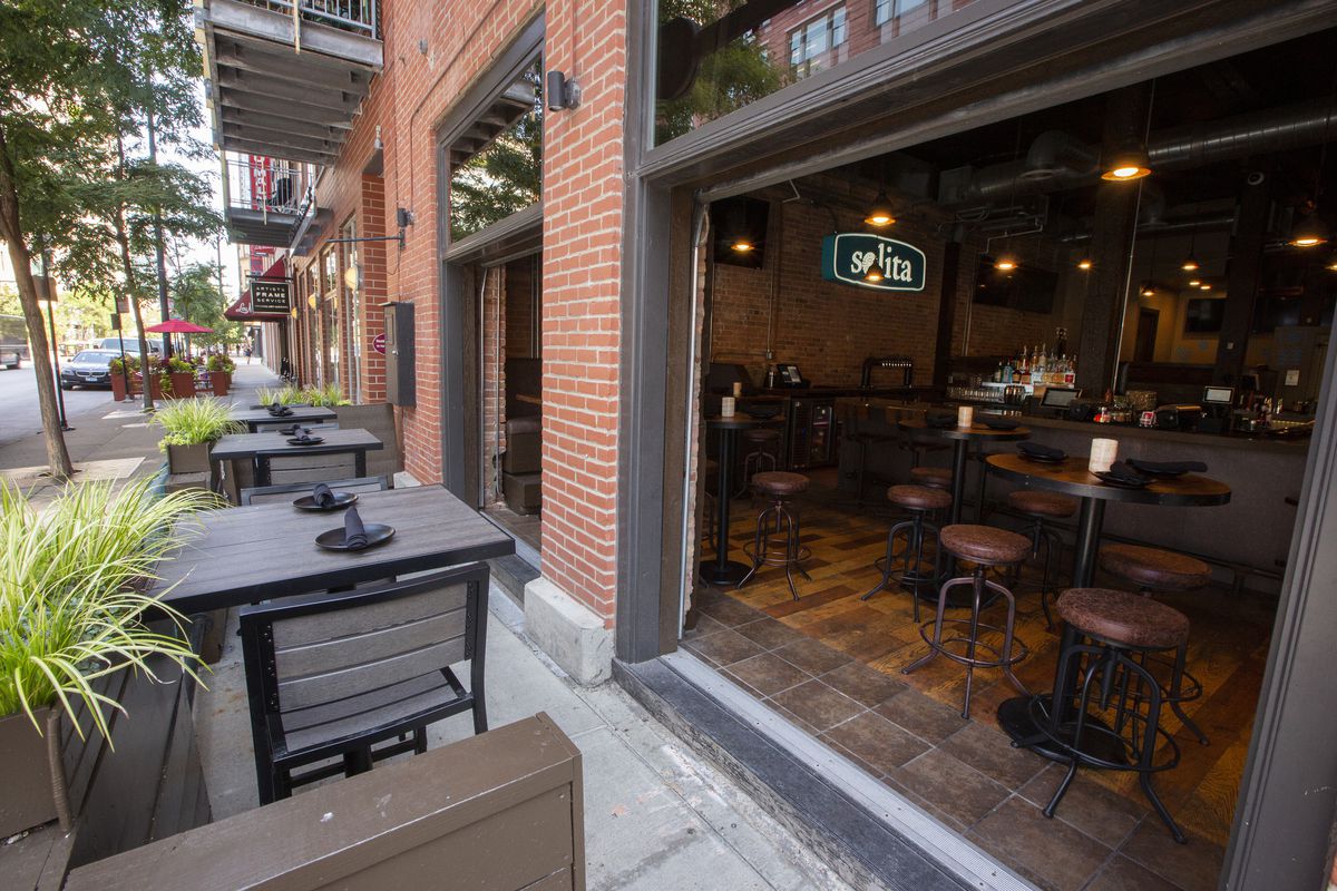 A large bar and restaurant space opens directly onto a narrow sidewalk patio along a tree-lined street.