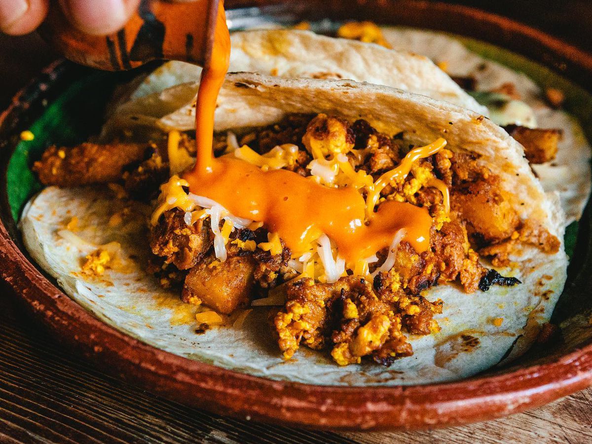 A hand pouring an orange sauce onto an open-face taco with brown-orange chunks, eggs, and cheese.