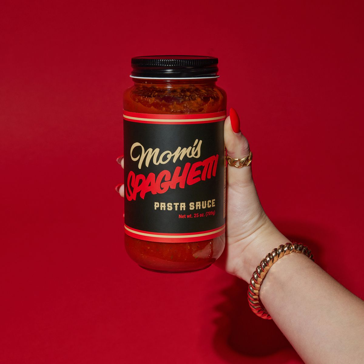 A hand with a red coffin nail on the thumb, a gold ring, and a gold bracelet on the wrist holding a jar of red sauce that says Mom’s Spaghetti Pasta Sauce.