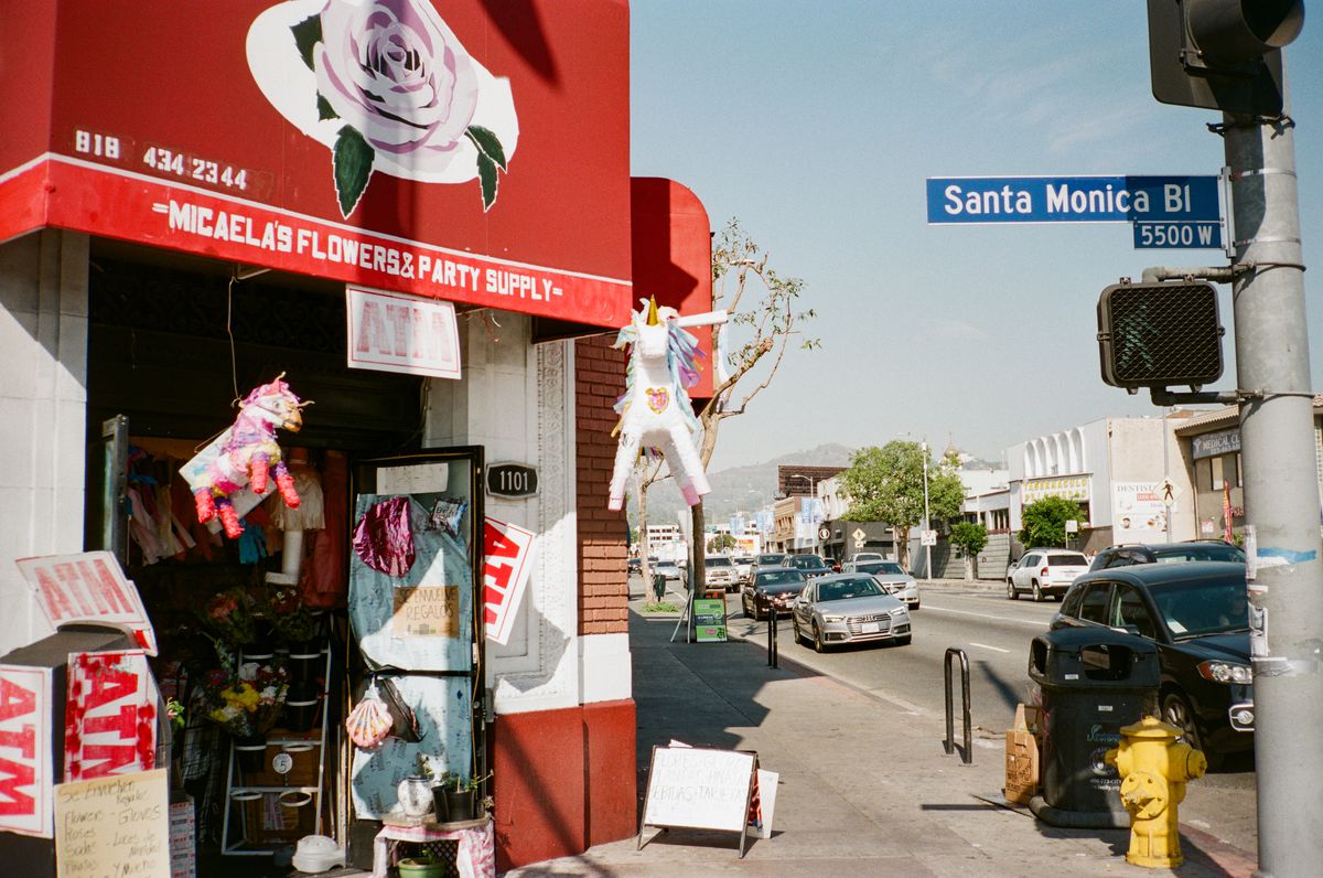 A red awning with a pink rose on it, the outside of Micaela’s Flowers &amp; Party Supply, has two unicorn pinatas hanging. The shop is at the corner of Santa Monica Blvd.