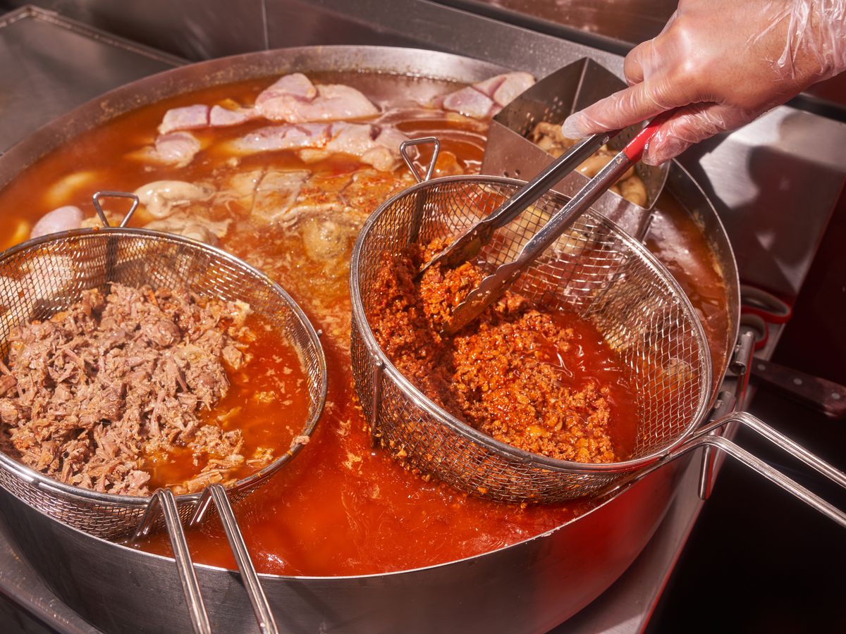 A gloved hands hold a sieve of crumbly red meat over a vat of orange fat and oil, also filled with other meats