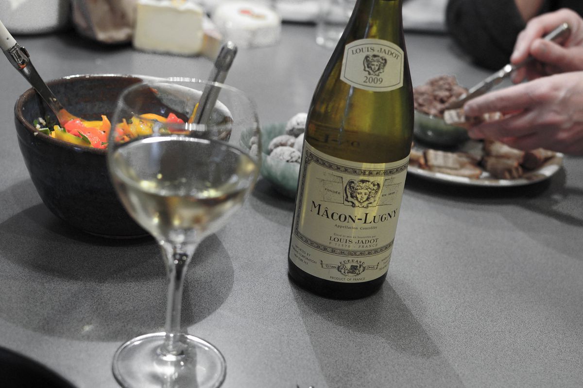 A bottle of white wine, with a filled glass, on a table surrounded by French food.