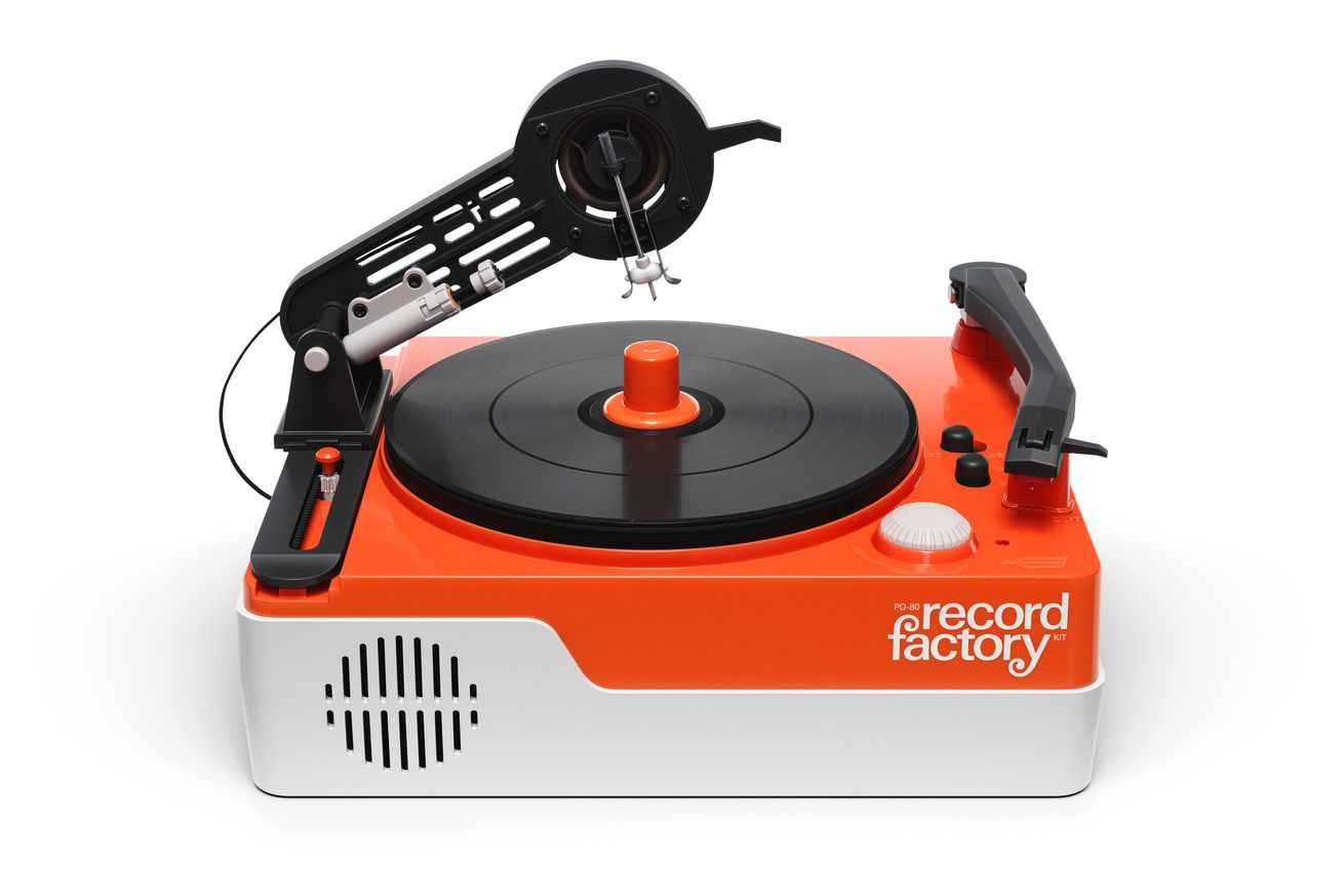The record factory is in a white background, the device is orange and black plastic with a black turntable on top, a speaker grille carve on the side, a resting playback arm, and a floating cutting arm that has multiple carving blades on it.