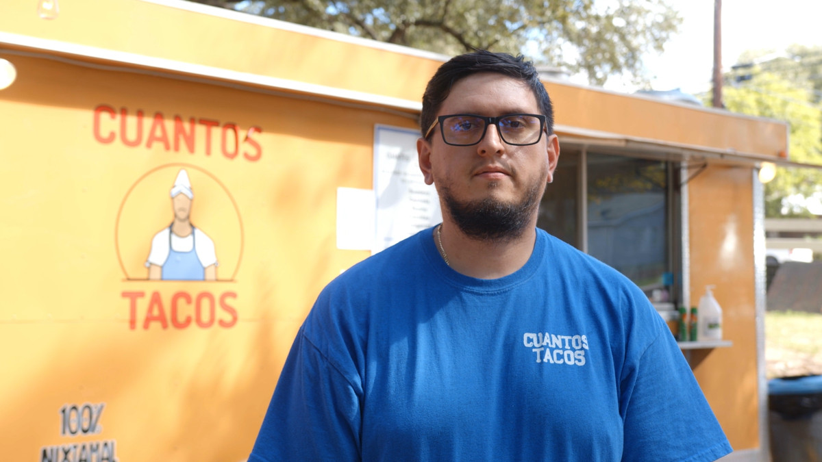 A man in a bright blue t-shirt and black-frame glasses in front of a yellow food truck with a sign reading “Cuantos Tacos.”