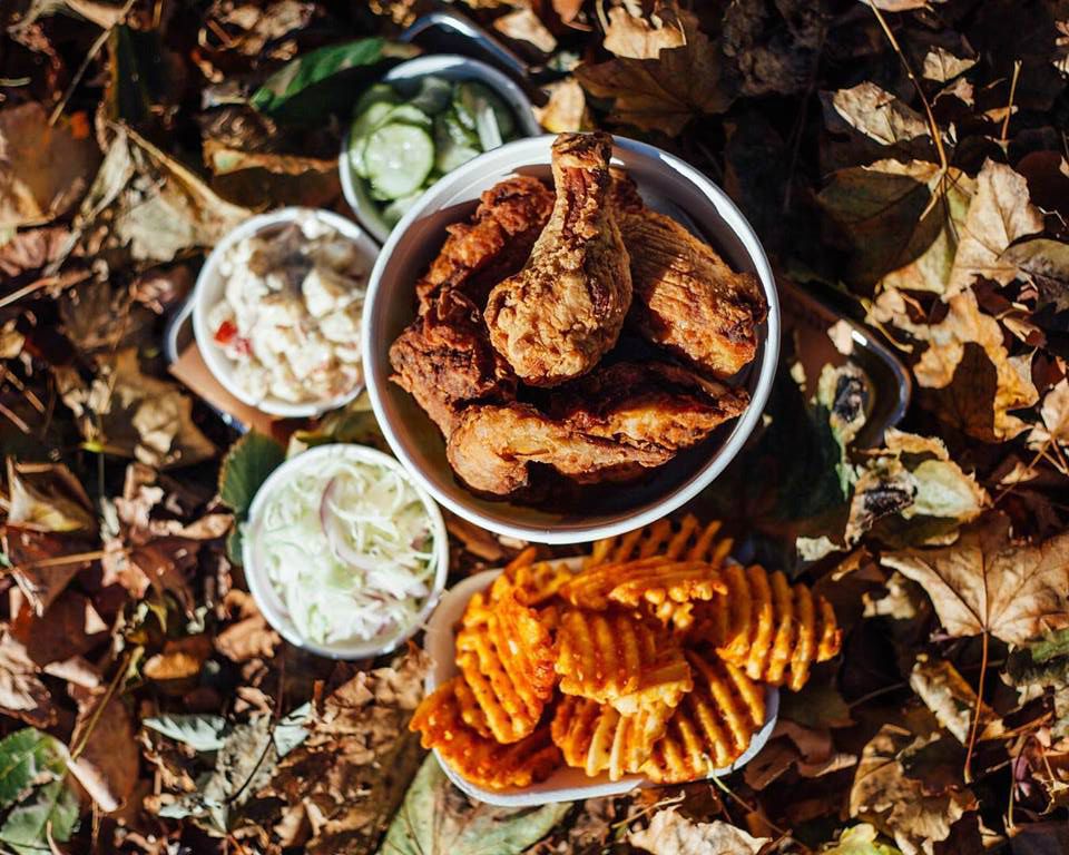 A bucket of fried chicken from Bucktown Chicken &amp; Fish sits on the ground, which is covered with fallen leaves. There are also waffle fries and other sides.