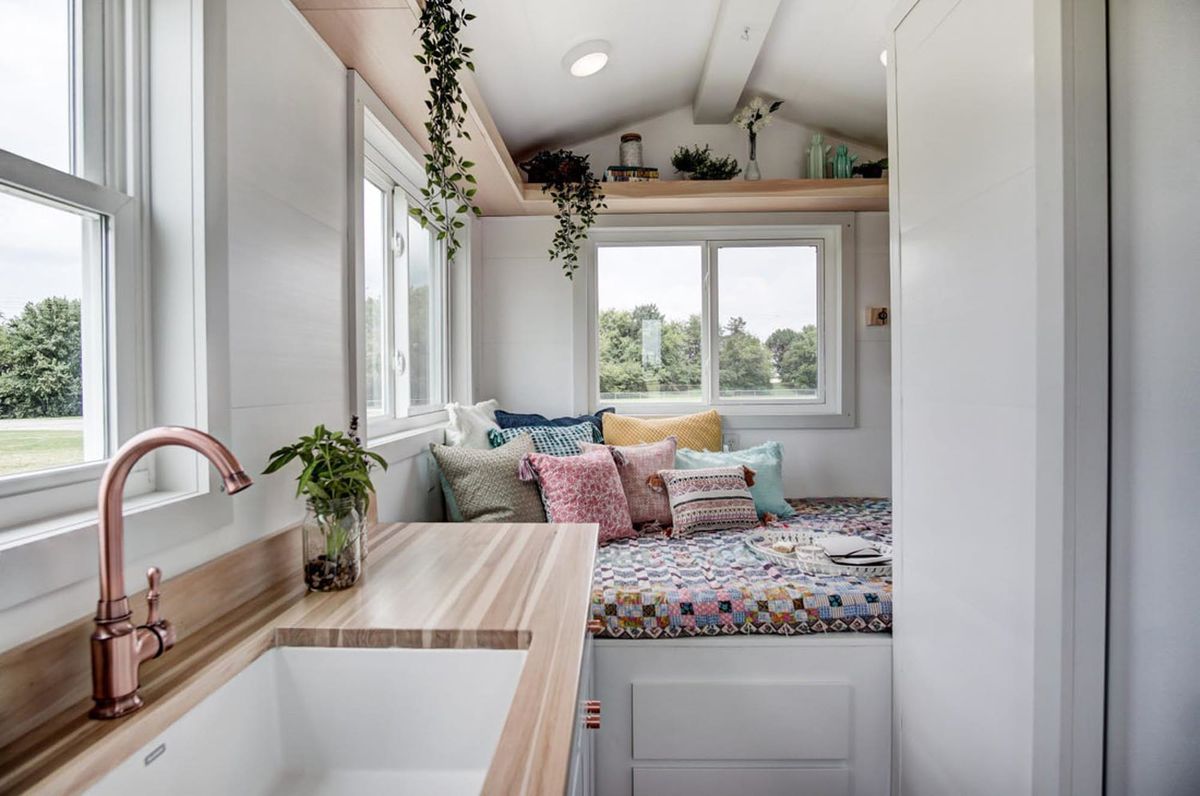 The interior of a tiny house. There is a sink, a bed with patterned bed linens and pillows, a shelf above the bed with assorted plants, and storage space under the bed. 