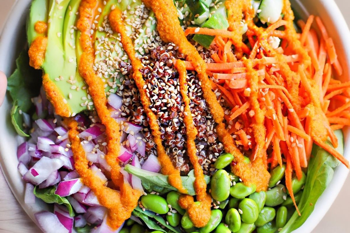 A salad bowl with carrots, avocado, edamame, chopped onion, red quinoa, and cucumbers is topped with an orange colored dressing. 