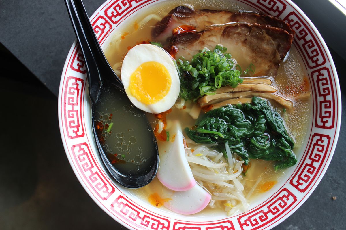 A bowl of ramen comes with a half of a boiled egg, mushrooms, and spinach, with a golden yellow broth