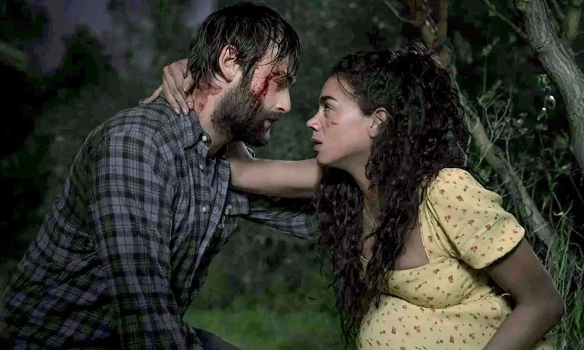 A pregnant Hanna John-Kamen wearing a yellow polka dot dress rests against a tree and puts her right arm around Douglas Booth, who has a bloodied face, in Unwelcome.