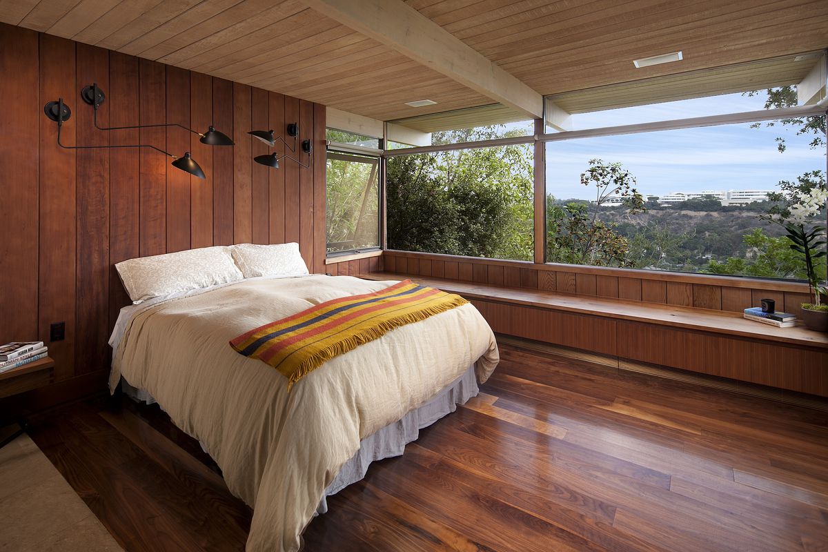 The master bedroom has wood paneled walls, wood floors, and wood beamed ceilings A bed sheathed in white linen abuts the wall.