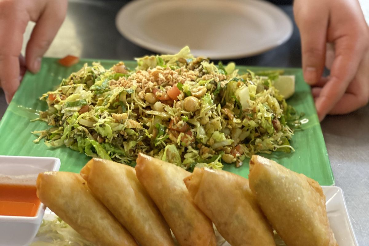 A white plate with five samosas sits next to a white  container of dipping sauce. A green plate of a chopped, slaw-like salad sits behind it, being placed by two hands