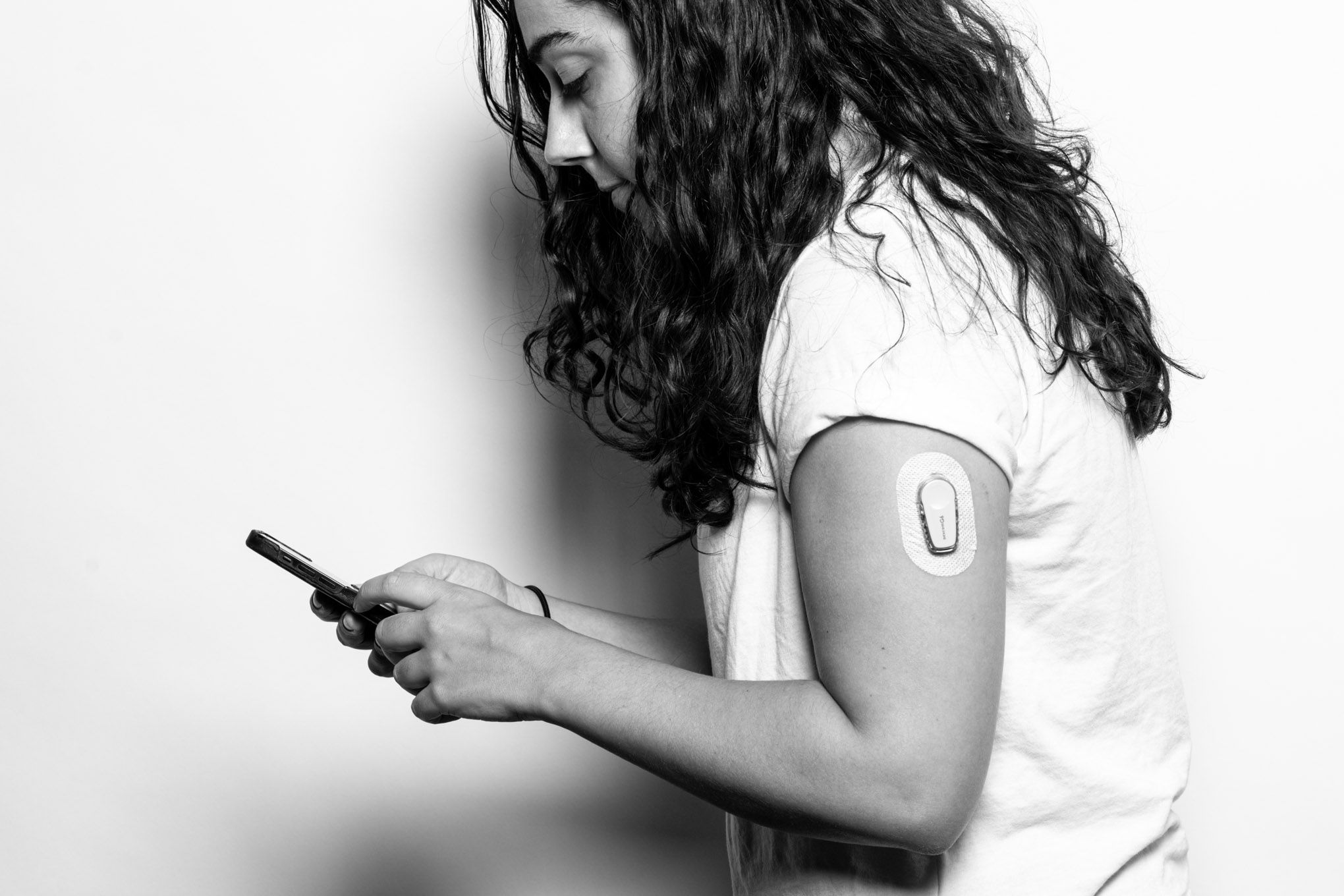 Une photographie en noir et blanc de Nicole regardant son téléphone.  Elle porte un glucomètre sur son bras supérieur gauche. 