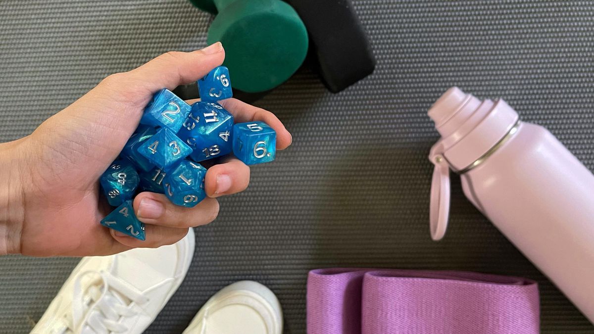 A hand holding some blue D&amp;D dice over an assortment of workout equipment, including two dumbbells, a water bottle, booty bands, and white sneakers