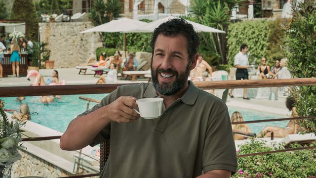Adam Sandler sits at an outdoor table holding up a mug and grinning in Hustle