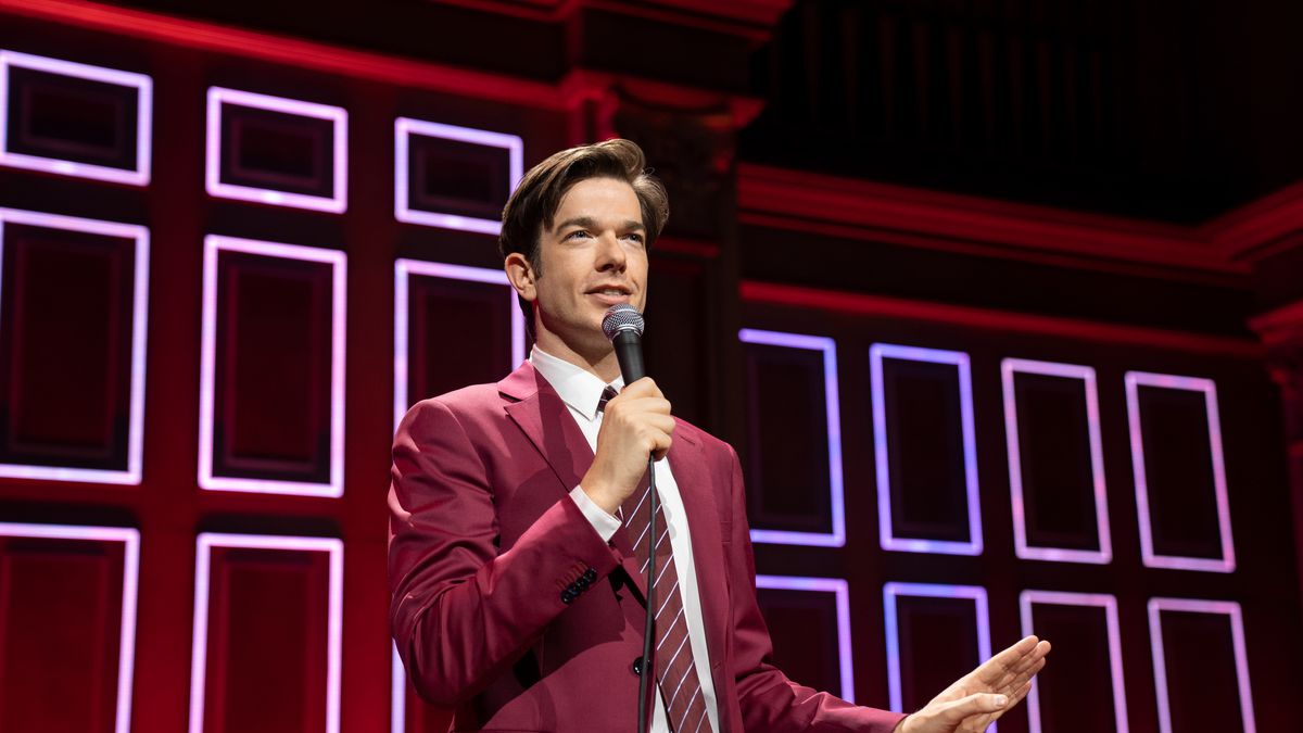 Comedian John Mulaney stands on stage with a maroon background complementing his red suit in a mid shot as he holds a mic to his face with a hand out in front of him in the Netflix special Baby J.