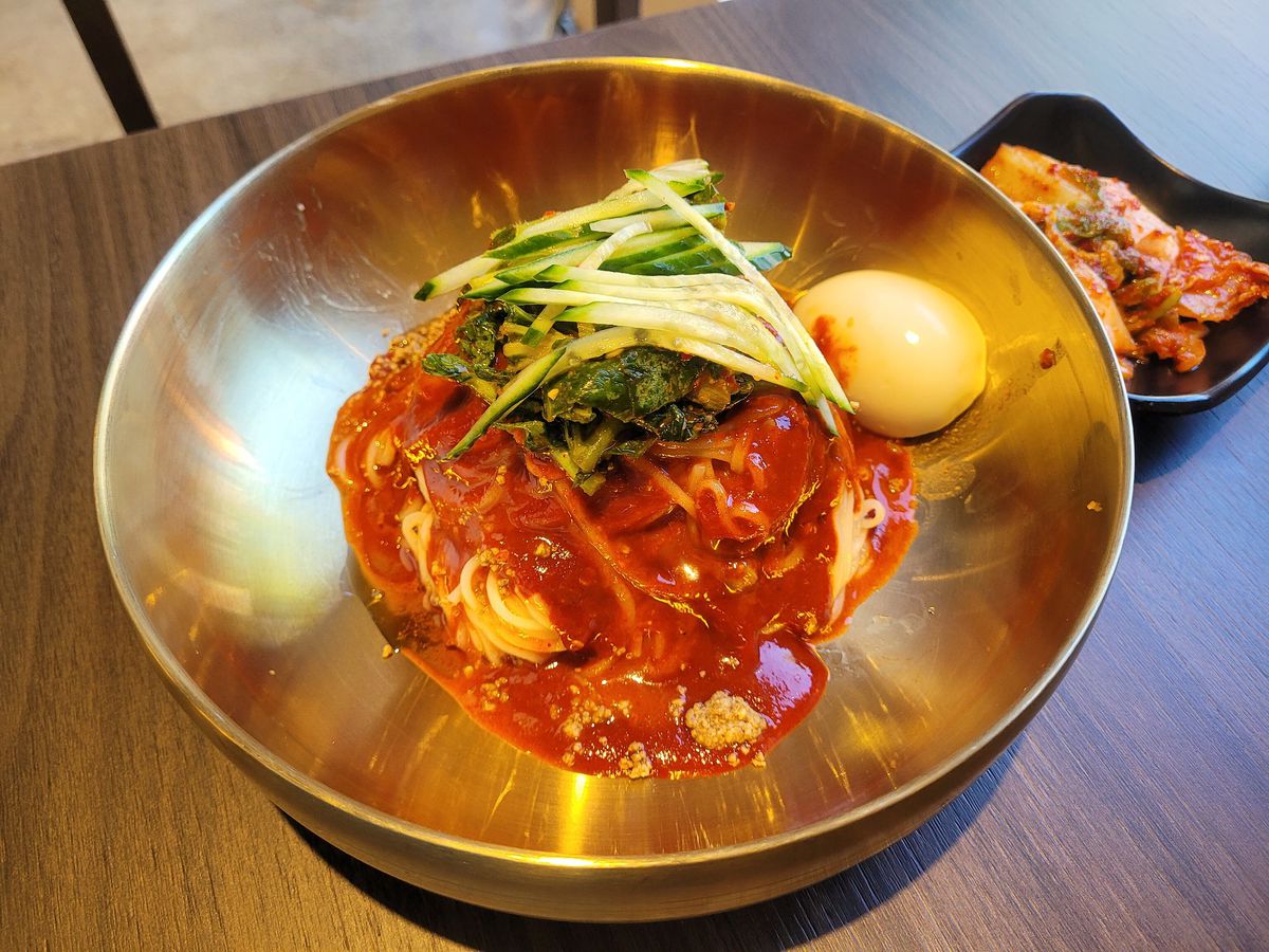 A knot of cold wheat noodles dressed in a fiery red sweet, spicy, sour chili sauce, topped with julienned cucumber and pickled greens with a side of half a boiled egg.