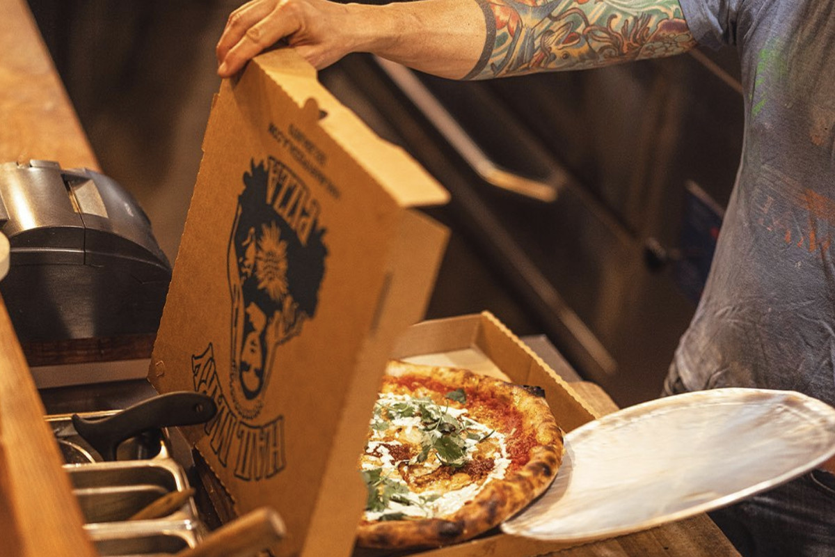 A chef with tattooed hands moves a pizza from a plate into a takeaway box.